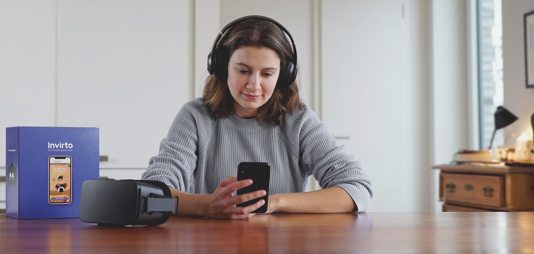 Eine Frau mit Kopfhörern sitzt am Tisch und schaut auf ihr Smartphone, neben ihr liegt eine VR-Brille