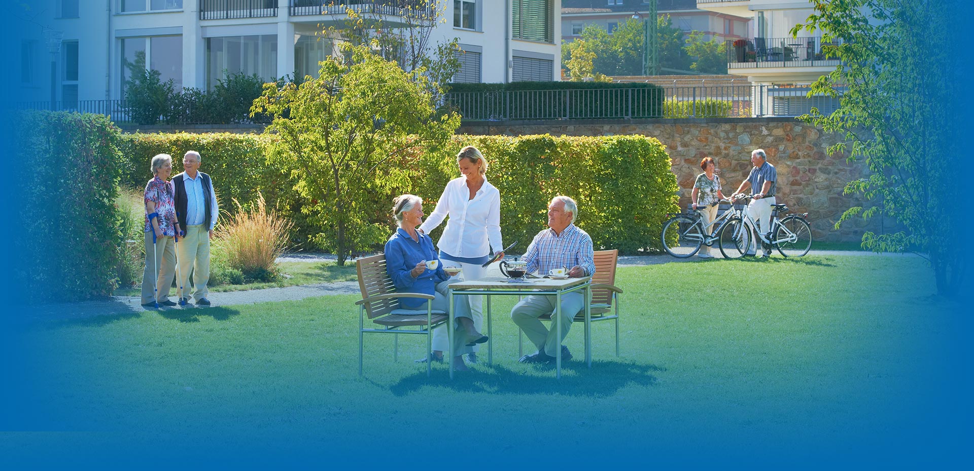 Szene im Garten vor betreutem Wohnen: Frau mit Krücken und Senior unterhalten sich, älteres Paar sitzt am Tisch, trinkt Kaffee und unterhält sich mit Pflegekraft, im Hintergrund älteres Paar mit Fahrrädern