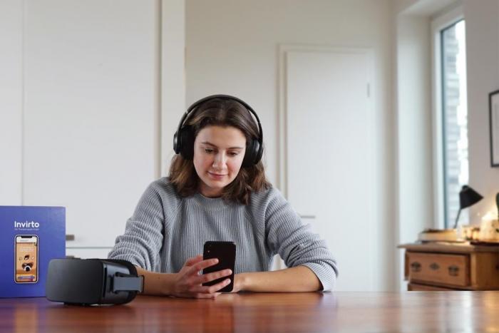Eine Frau mit Kopfhörern sitzt am Tisch und schaut auf ihr Smartphone, neben ihr liegt eine VR-Brille